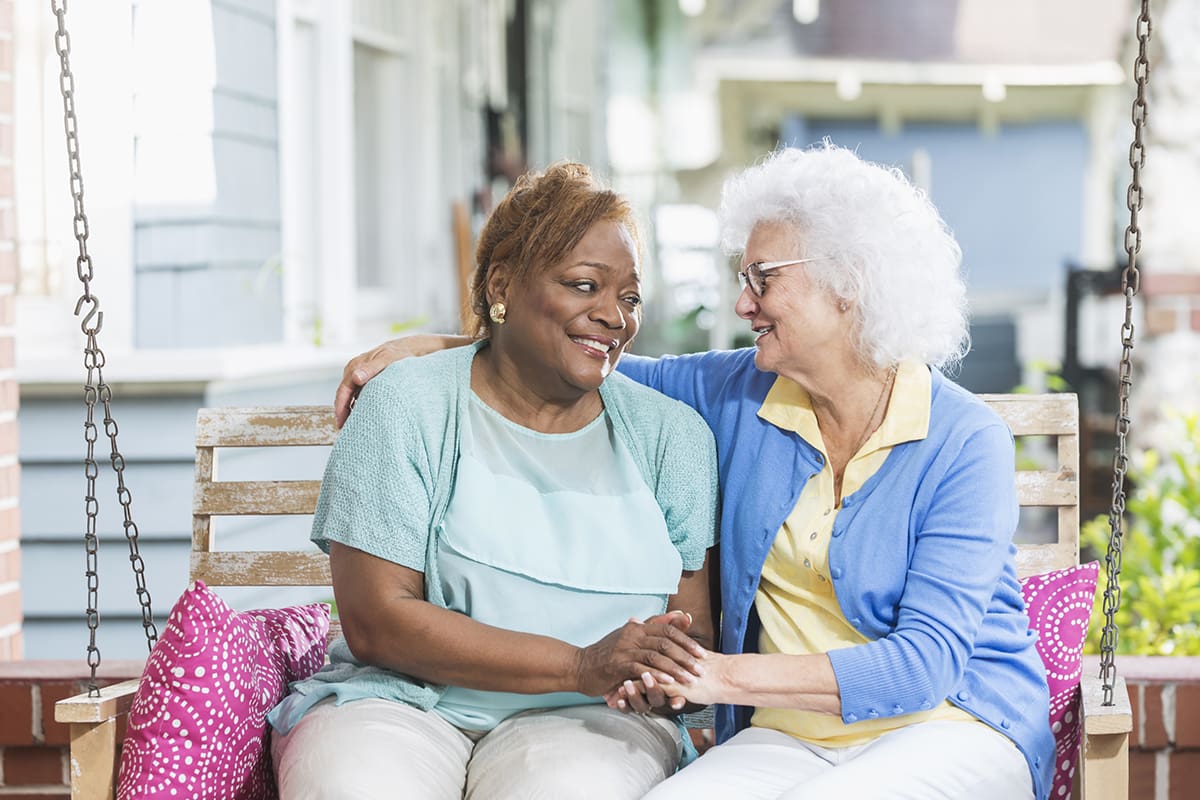 Two multi-ethnic senior woman, best friends, sitting together on a porch swing, smiling and looking at each other. They are relaxed. The older woman, in her 70s with white hair, has her arm around the shoulder of her African American friend.